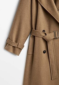 Manteau en laine marron avec larges revers, boutons noirs croisés, taille ceinturée et détail de sangle au poignet de la manche.
