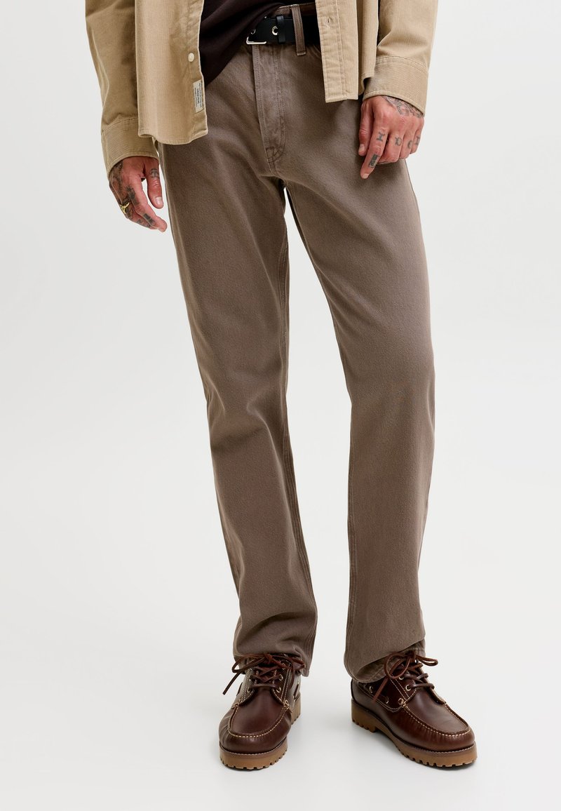 Homme portant un pantalon marron à jambes droites, des chaussures en cuir foncé à lacets, une ceinture noire et une chemise beige avec des tatouages visibles sur les mains.
