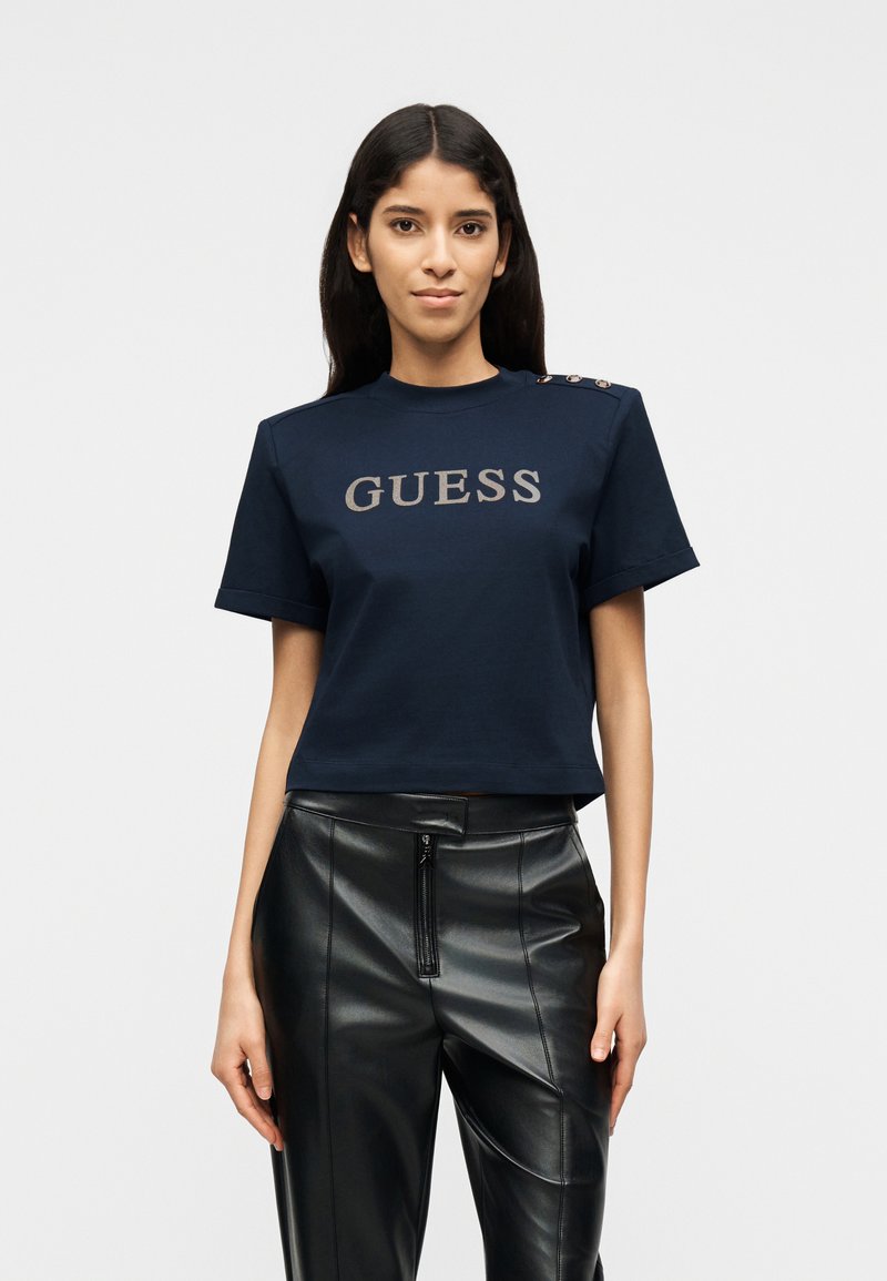 Γυναίκα που φοράει μπλε ναυτικό t-shirt GUESS με κουμπιά στον ώμο και μαύρο ψηλόμεσο παντελόνι από τεχνητό δέρμα.