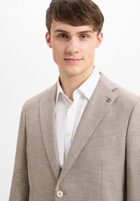 Helles beige Blazer aus strukturiertem Stoff, mit einem einzelnen Knopfverschluss und einem dezenten Logo am Kragen, getragen über einem weißen Hemd.