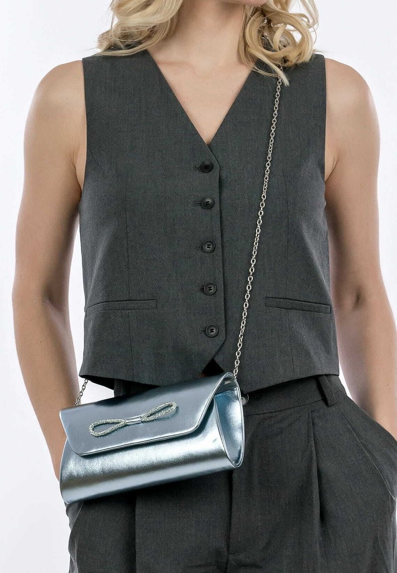 Femme portant un gilet sans manches gris foncé boutonné et un pantalon assorti, tenant une pochette bleue métallique brillante avec une bandoulière en chaîne et un nœud décoratif.