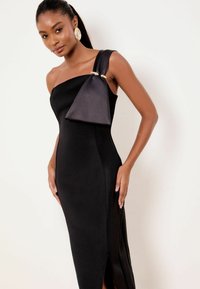 Robe noire moulante à une épaule avec un accent triangulaire en satin, une garniture dorée et des fentes latérales pour plus de détails. Texture lisse.