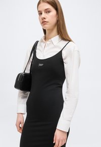 Νέα γυναίκα που φοράει μαύρο αμάνικο φόρεμα Tommy Jeans πάνω από λευκό πουκάμισο με κουμπιά, κρατώντας μια μικρή μαύρη τσάντα ώμου.