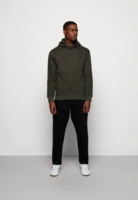 Olivzaļš kapučjakas džemperis ar kenguru kabatu, ar faktūru audumu, kopā ar melnām pieguļošām biksēm un baltām sporta kurpēm. Vienkāršs, sportisks dizains.