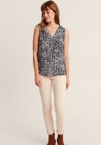 Top senza maniche a scollo a V con motivo di foglie blu scuro e crema; abbinato a pantaloni attillati beige chiaro e stivali alla caviglia marroni.