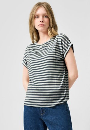 Junge Frau mit schulterlangen blonden Haaren, die ein grau-schwarz gestreiftes Kurzarmshirt und blaue Jeans trägt, stehend mit den Händen hinter dem Rücken.