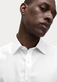Camisa blanca de botones con cuello puntiagudo, textura suave y un bolsillo en el pecho. Presenta botones simples y redondeados en la parte delantera.