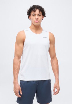 Joven con pelo rizado que lleva una camiseta deportiva blanca sin mangas y pantalones cortos azul marino, de pie contra un fondo claro y liso.