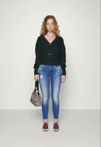Cardigã canelado verde escuro com três botões, combinado com jeans skinny desfiados azul claro, sapatos em estampa de leopardo rosa e uma bolsa em estampa de zebra preto e branco.