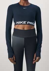 Henkilö, joka on pukeutunut mustaan pitkänhihaiseen lyhyttoppiseen urheiluvaatteeseen, jossa on "NIKE PRO" -nauha, sekä mustiin korkeavyötäröisiin legginsseihin, seisoo yksinkertaista taustaa vasten.