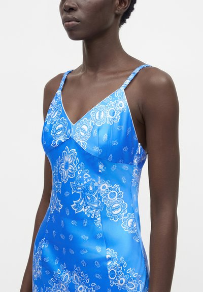 Femme portant une robe bleue sans manches avec des motifs floraux blancs et cachemire, avec de fines bretelles froncées.