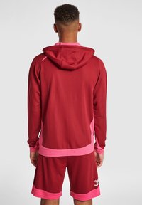 Felpa sportiva rossa con dettagli rosa, realizzata in un tessuto strutturato, con cappuccio e polsini a coste. Abbinata a pantaloni corti rossi coordinati.