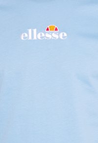 Světle modré tričko z bavlněné tkaniny, s bílým vyšitým logem "ellesse" a barevným akcentem loga nad ním.