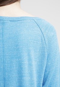 Ljusblått, texturerat tyg med bred halsringning och raglanärmar, med ett subtilt melerat mönster och sömdetaljer på baksidan.