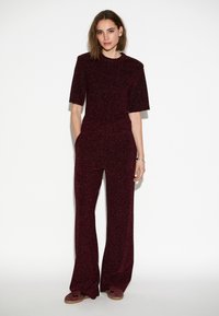 Bordeaux gebreide jumpsuit met korte mouwen, wijde benen, textuurstof en zijzakken. Model staat tegen een effen achtergrond.