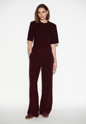 Bordeaux gebreide jumpsuit met korte mouwen, wijde benen, textuurstof en zijzakken. Model staat tegen een effen achtergrond.