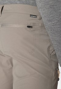 Beige lauko kelnės, pagamintos iš elastingos medžiagos, su šonine kišene su užtrauktuku, plokščiomis siūlėmis ir logotipo etikete ant juosmens.