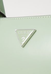 Green textured handbag featuring a triangular silver logo with "GUESS U.S.A." and a question mark, with contrast stitching along the edges.