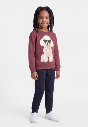 Fille aux cheveux tressés portant un sweat-shirt bordeaux avec un motif caniche, un pantalon bleu marine et des baskets blanches, debout devant un fond uni.