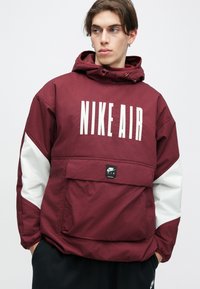 Camisola com capuz Nike Air numa cor bordeaux com realces brancos, de corte oversized, com bolso frontal e texto do logótipo em destaque; o tecido parece suave e durável.