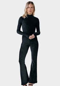 Woman with blonde hair wearing a fitted black long-sleeve top and matching flared pants, standing with hands clasped.