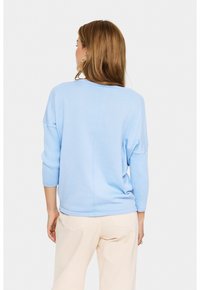 Donna con capelli castani che indossa un maglione a maniche lunghe azzurro chiaro e pantaloni beige, in piedi con le spalle rivolte verso di noi su uno sfondo bianco.