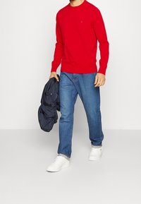 Rode gebreide trui, blauwe denim jeans, witte sneakers; houdt een marineblauwe lichtgewicht jas vast. Heeft een klein logo op de trui.