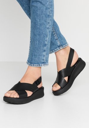 Füße in schwarzen Plattformsandalen mit überkreuzten Riemen und Knöchelriemen, kombiniert mit blauen Jeans, die am Knöchel gekürzt sind.