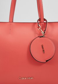 Sac cabas corail en cuir texturé, doté de deux anses et d'une petite pochette ronde amovible pour les pièces. Logo doré sur le devant.