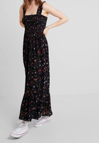 Robe longue en tissu noir à motifs floraux rouges, bleus et jaunes. Corsage smocké et bretelles à volants ; portée avec des baskets blanches.