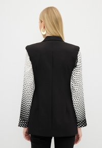 Blazer noir avec des épaules structurées, présentant des manches blanches à motifs qui passent de points à du noir uni. Texture lisse, coupe ajustée.