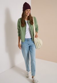 Jeune femme portant une veste verte, une chemise blanche, un jean bleu clair, des baskets blanches et un bonnet violet, portant un sac beige à l'épaule.