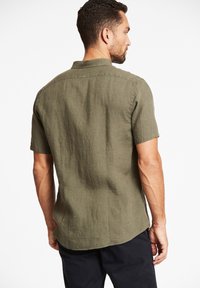 Korte mouwen, olijfgroen shirt van gestructureerde stof; voorzien van een standaardkraag en een geschulpte zoom. Zichtbare stiksels op de naden.