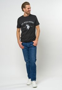 Sort bomulds T-shirt med hvid grafik af en polo-spiller, parret med blå denim jeans og hvide sneakers. Casual pasform, korte ærmer.