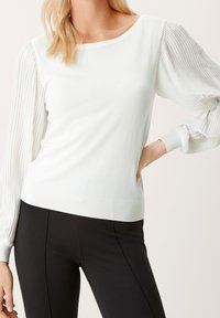 Pull blanc avec des manches plissées et bouffantes et un ourlet côtelé, associé à un pantalon noir ajusté. Le tissu semble lisse et léger.