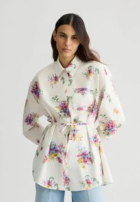 Cvećasta bluza s dugim rukavima, bijela osnova, višebojni uzorci cvijeća, prednji džep ukrašen rhinestonesom i pojas oko struka.
