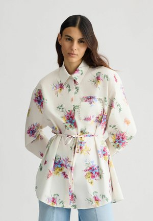 Floral blouse with long sleeves, white base, multicolored flower prints, a front pocket adorned with rhinestones, and a waist tie.