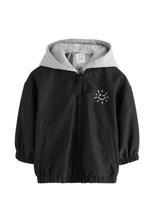 Veste coupe-vent noire avec capuche grise, poignets élastiques et fermeture éclair. Présente un petit graphique de soleil sur la poitrine. Texture douce et lisse.