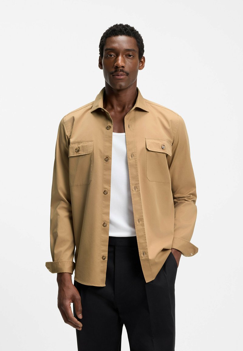 Homme aux cheveux courts et bouclés avec une moustache, portant une chemise marron clair boutonnée sur un débardeur blanc et un pantalon noir, debout devant un fond blanc.