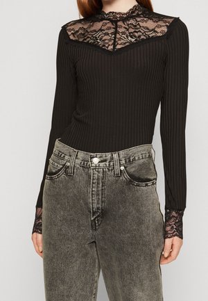 Sort ribstrikket bluse med lange ærmer, blondeindlæg og manchetter, kombineret med højtsiddende, falmet sorte jeans på en person.