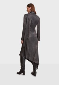 Dlhé čierne šaty s vysokým golierom, asymetrickým lemom a hladkým, jemne textúrovaným materiálom; v kombinácii s čiernymi čižmami po kolená.