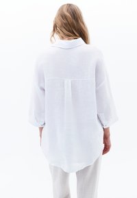 White, lightweight linen shirt with a back pleat, buttoned cuffs, and a curved hem. Features a classic collar and relaxed fit.