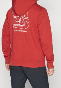 Roter Hoodie mit einer Kordelzugkapuze, auf der Rückseite mit einem weißen Grafikdruck, der "SIND FÜR DAS KLETTERN BESTIMMT" besagt. Weiches Material, gerippte Bündchen.
