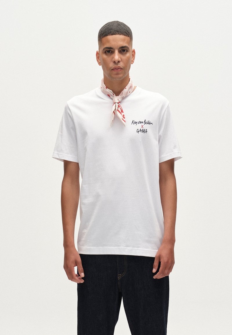 Tricou alb din bumbac cu mâneci scurte, având text negru pe pieptul stâng. Eșarfă cu model roșu și alb legată la gât. Blugi închiși la culoare.