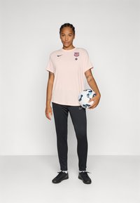 Svijetlo ružičasta Nike majica s logotipom FC Barcelone, uparena s crnim sportskim hlačama i crnim sportskim cipelama, drži bijelu nogometnu loptu.
