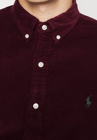 Camisa de botón de pana burdeos con cuello clásico, botones blancos y un sutil logo verde bordado en el pecho.