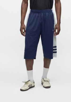 Homme portant un long short de basket bleu marine avec des panneaux latéraux gris et des baskets blanches avec des rayures noires, debout sur fond blanc.