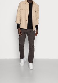 Beige katoenen jas met voorzakken, gecombineerd met een zwarte trui en donkere cargobroek, afgewerkt met witte sneakers. Eenvoudig ontwerp en texturen.