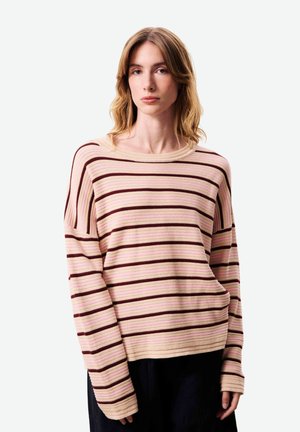 Femme aux cheveux châtains mi-longs portant un pull à rayures beige, rose et marron foncé et un pantalon noir.