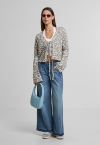 Zkrácený, vícebarevný pletený svetr s dlouhými rukávy, spojený s širokými modrými džíny. Drží světle modrou, texturovanou kabelku.